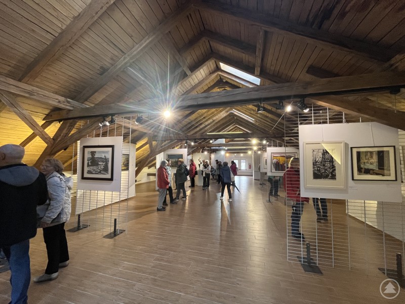 Besucherinnen und Besucher schlendern durch eine helle Dachgalerie mit Holzbalkendecke, in der zahlreiche Kunstwerke ausgestellt sind.