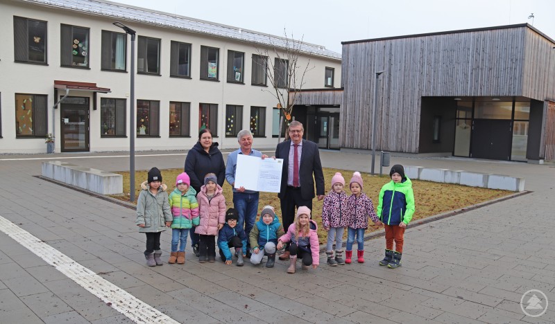 Hans-Peter Schmucker, Leiter des ALE Niederbayern (rechts), überreichte an Bürgermeister Leo Meier (Mitte) die Auszahlungsurkunde. Mit dabei waren Kindergartenkinder zusammen mit Leiterin Susanne Steininger. Gruppenfoto vor dem neu gestalteten Dorfgemeinschaftshaus in Kumreut. Zu sehen sind Bürgermeister Leo Meier, der Leiter des ALE Niederbayern Hans-Peter Schmucker sowie mehrere Kindergartenkinder mit ihrer Leiterin vor dem Gebäudeensemble aus Altbau und moderner Halle.