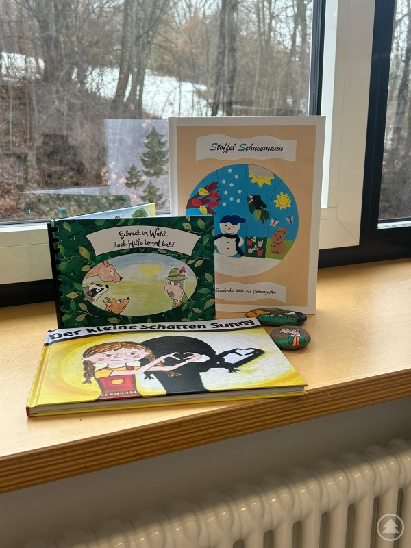 Die im W Seminar entstandenen Kinderbücher bilden die Grundlage für das LLG Lesemobil auf Tour. Mehrere selbst gestaltete Kinderbücher und bemalte Steine stehen auf einer Fensterbank. Im Hintergrund ist eine winterliche Landschaft vor dem Fenster zu sehen.