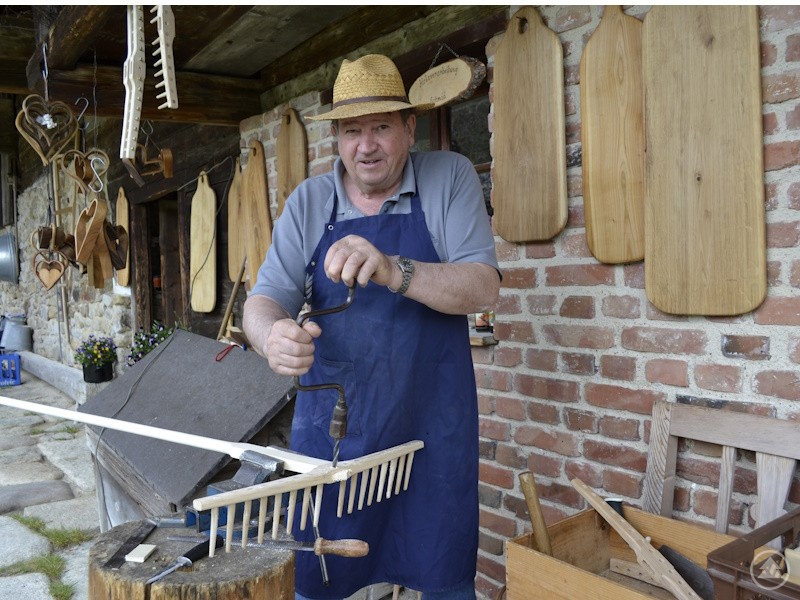 Max Schmid zeigt im Freilichtmuseum Finsterau, wie hölzerne Rechen traditionell per Hand gefertigt werden Ein Handwerker mit Strohhut und blauer Schürze arbeitet an einem hölzernen Rechen vor einer rustikalen Steinwand im Freilichtmuseum Finsterau.