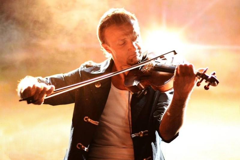 Ein Mann mit offener Jacke und weißem Shirt spielt konzentriert Geige vor einem leuchtenden Hintergrund.