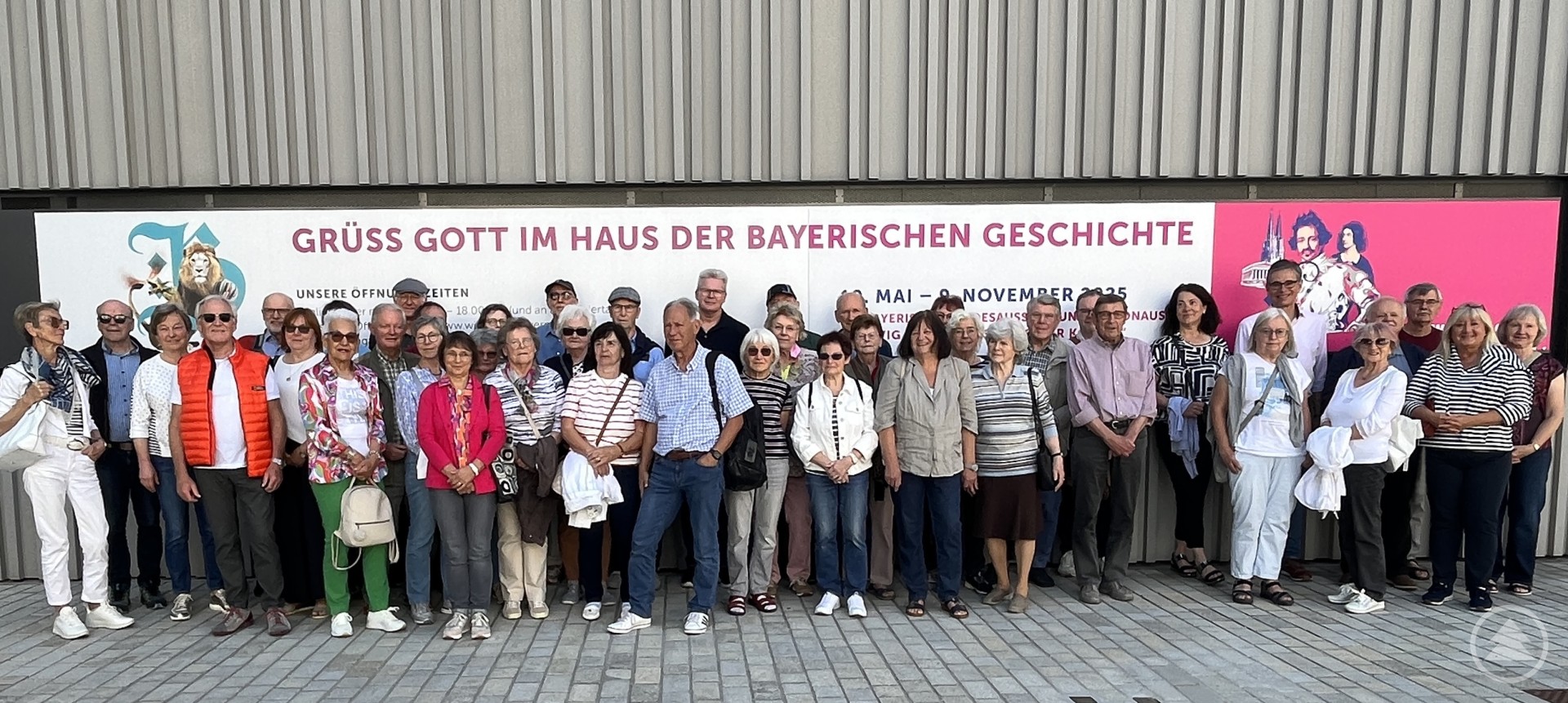 Eine große Gruppe älterer Menschen steht dicht beieinander für ein Gruppenfoto vor dem „Haus der Bayerischen Geschichte“. Im Hintergrund ist ein großes Plakat zur Ausstellung „Ludwig I.“ zu sehen.