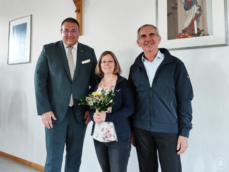 Ein Blumengruß für die neue Pfarrgemeinderätin: Stadtpfarrer Magnus König (re.) und Pfarrgemeinderatsvorsitzender Christoph Weishäupl (li.) heißen Marion Schlutz im Gremium willkommen. Drei Personen stehen nebeneinander vor einer weißen Wand mit religiösen Bildern. Die Frau in der Mitte hält einen Blumenstrauß, die beiden Männer links und rechts lächeln.