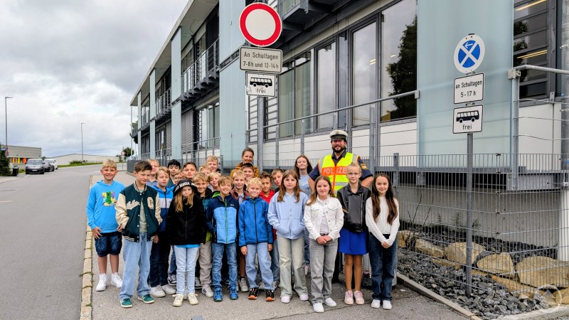 Eine Gruppe von Schülerinnen und Schülern steht mit einem Polizisten in Warnweste und einer Lehrkraft vor der Realschule Grafenau, im Hintergrund Verkehrsschilder.