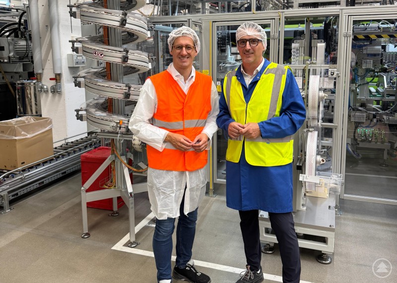 Zwei Männer mit Schutzkleidung, Sicherheitswesten und Haarnetzen stehen in einer modernen Produktionshalle vor Maschinen und Förderanlagen.