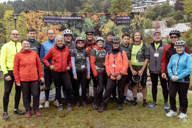 Zum Auftakt des „5-Gänge-Radelns“ starteten die Teilnehmer gemeinsam am Wanderparkplatz in Bayerisch Eisenstein. Eine große Gruppe Radfahrer in bunter Sportkleidung posiert vor einem Banner mit der Aufschrift „Ferienregion Nationalpark Bayerischer Wald“.