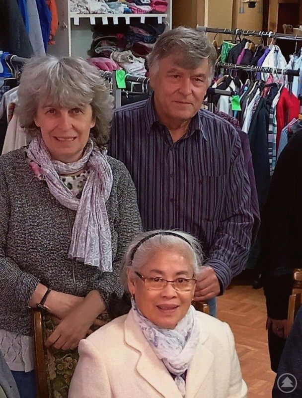 Drei Personen stehen bzw. sitzen in einem Raum mit Kleiderständern und Regalen voller Kleidung. Sie lächeln in die Kamera und wirken herzlich und engagiert.