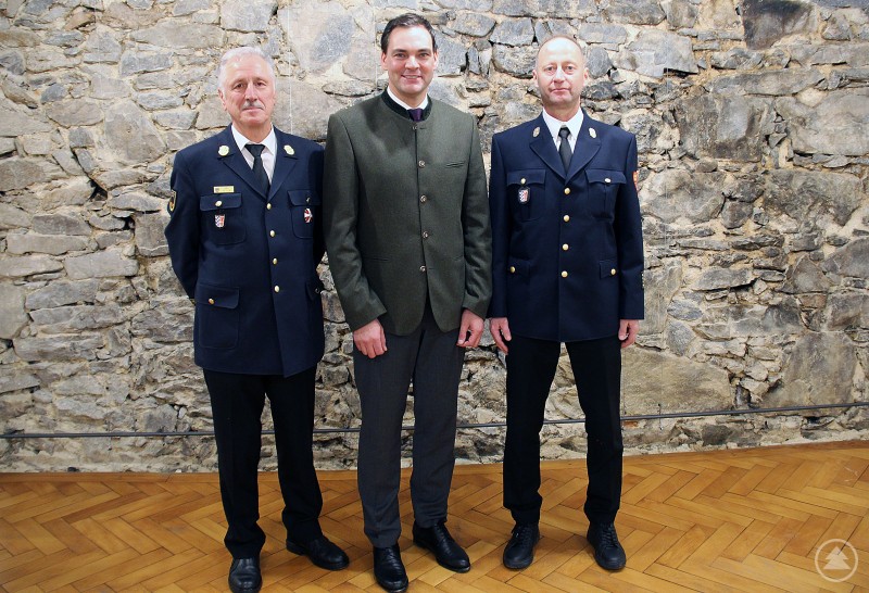 v.l. Ehrenkreisbrandrat Norbert Süß, Landrat Sebastian Gruber und der neue Kreisbrandrat Martin Bauer Drei Männer stehen in einem feierlichen Rahmen vor einer Steinwand. Zwei von ihnen tragen Feuerwehr-Uniform, der Dritte steht in der Mitte im Trachtenanzug. Alle blicken freundlich in die Kamera.