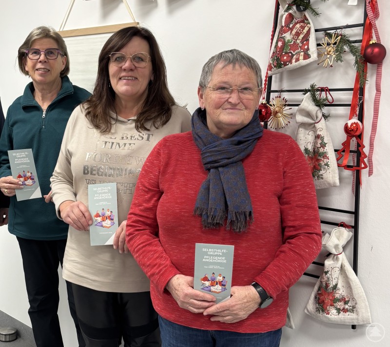 Die Leiterinnen der Selbsthilfegruppe (v. l.) Christl Großer, Heidi Töpfl und Renate Kloiber wollen auch 2026 praktische Tipps an die Hand geben und vor allem ein Gefühl der Solidarität und gegenseitigen Unterstützung vermitteln. Drei Frauen stehen nebeneinander in einem Innenraum und halten jeweils eine Broschüre in den Händen. Hinter ihnen ist eine dekorierte Wand mit Taschen und Textilien zu sehen. Es handelt sich um die Leiterinnen der Selbsthilfegruppe für pflegende Angehörige im Landkreis Freyung Grafenau.