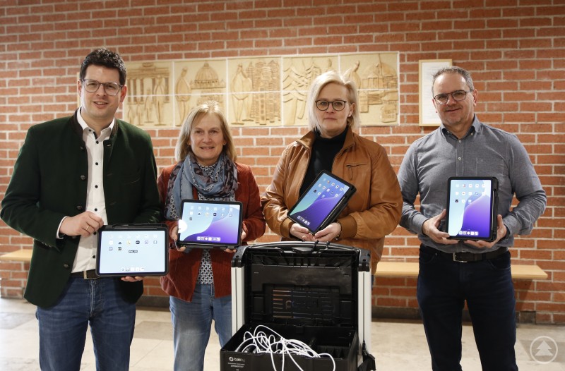 Bürgermeister Martin Pichler, Rektorin Roswitha Bumberger, Gemeinderätin Carolin Garhammer Referentin Schule und kommunaler IT Fachmann Robert Meier sind froh, dass 18 weitere Tablets Schule und Lernen ergänzen und unterstützen. Vier Personen stehen vor einer Backsteinwand und präsentieren neue Tablets für die Schule. Vor ihnen befindet sich ein Transportkoffer mit Ladefunktion für mehrere Geräte.