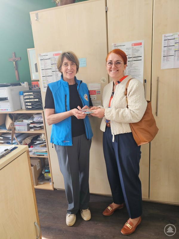 v.l.: Angelika Leitl-Weber, Leiterin der Bahnhofsmission, und Andrea Kleingütl, stellv. Leitung Personal vom Bistum Passau bei der Übergabe der Spende aus der Aktion „Blumen kaufen und Gutes tun“ Zwei Frauen stehen in einem Büro vor einem Schrank und übergeben sich einen Geldbetrag. Beide lächeln in die Kamera, im Hintergrund sind Unterlagen und ein Kreuz an der Wand zu sehen.