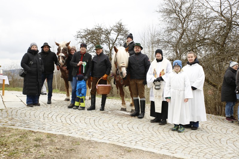 Erich Roth (mit Korb), Bürgermeister Martin Pichler (mit Erinnerungsanstecker) und Pfarrer Simon Steinbauer (mit Weihwasserkessel) ließen sich von Rosserern, Ministranten, dem Mesnerteam und vielen weiteren Helfern von Dorf und Kommune unterstützen, um den Palmritt 2026 wieder zur Freude von Tier, Mensch und Zaungästen prächtig, erfolgreich und unfallfrei durchzuführen. Eine große Gruppe von Reitern, Helfern und Organisatoren steht gemeinsam mit Pferden für ein Gruppenfoto.