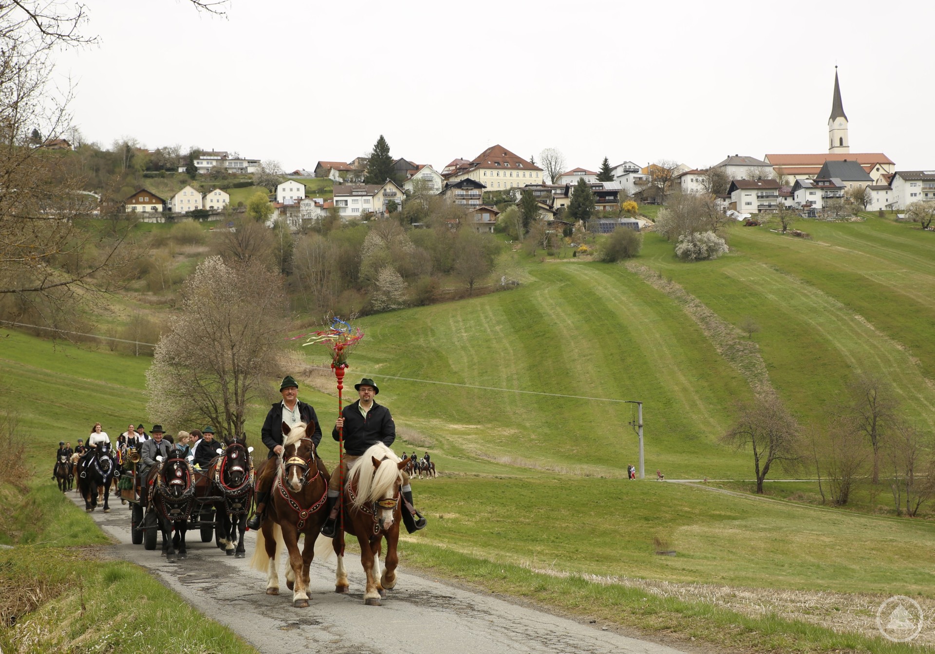 Angeführt vom Palmbuschen ging es von Schönberg nach Kirchberg hinüber. Das Bild zeigt einen festlichen Reiterzug auf dem Weg durch eine sanft hügelige Frühlingslandschaft. Angeführt wird der Palmritt von zwei Reitern auf prächtig geschmückten Pferden, einer davon trägt einen bunt geschmückten Palmbuschen. Dahinter folgen zahlreiche Teilnehmer mit Pferden und Kutschen.