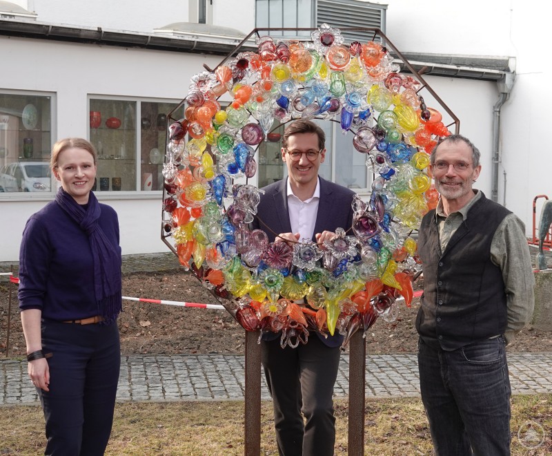 Wollen als "Botschafter des Werkstoffs Glas" Aufmerksamkeit für die Anliegen der Branche erzielen: (von links nach rechts) Die stellv. Schulleiterin der Glasfachschule Zwiesel Iris Haschek, MdL Dr. Stefan Ebner und der Schulleiter der Glasfachschule Zwiesel Gunther Fruth. Drei Personen stehen vor einem bunten Glaskunstwerk im Außenbereich der Glasfachschule Zwiesel und lächeln in die Kamera.