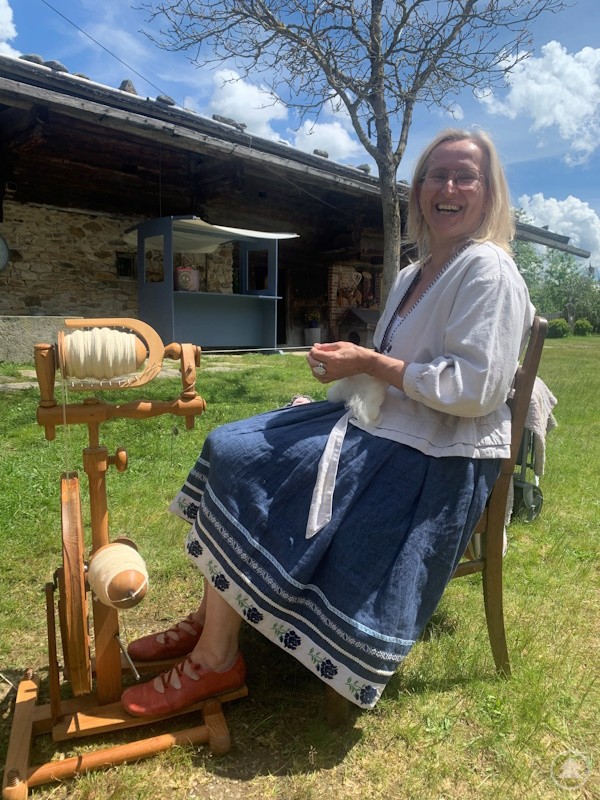 Eine Frau in traditioneller Kleidung sitzt vor einem Spinnrad und zeigt, wie Wolle zu Garn gesponnen wird. Im Hintergrund eine ländliche Kulisse.