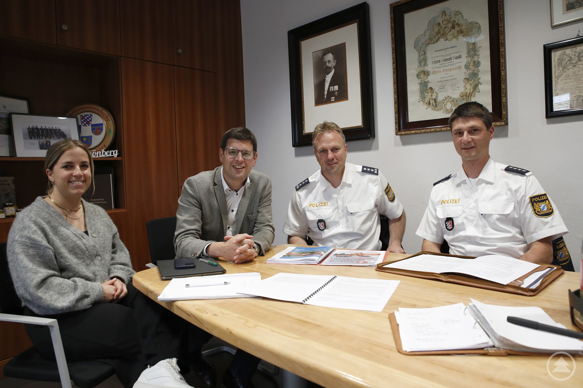 Zum Gespräch über Sicherheit im PI-Bereich Grafenau und im Markt Schönberg trafen sich vom Bürgerbüro Sophia Bumberger (v.l.), Bürgermeister Martin Pichler sowie die EPHK Thomas Kern und PHK Franz Weinzierl. Vier Personen sitzen gemeinsam an einem Besprechungstisch in einem Büro, zwei davon in Polizeiuniform, zwei in ziviler Kleidung. Auf dem Tisch liegen Akten und Unterlagen. Im Hintergrund hängen Urkunden und Fotos an der Wand.