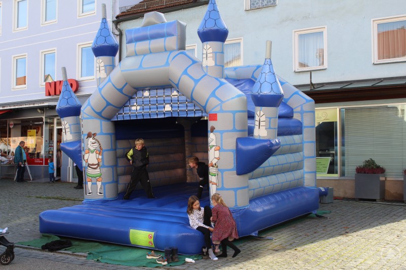 Die Hüpfburg hatte beim Aidenbacher Herbstmarkt magische Anziehungskraft auf die Kinder. Kinder toben fröhlich in einer großen blauen Hüpfburg in Form eines Märchenschlosses auf dem Marktplatz. Schuhe liegen am Rand, Passanten gehen vorbei, im Hintergrund sind Geschäfte zu sehen.