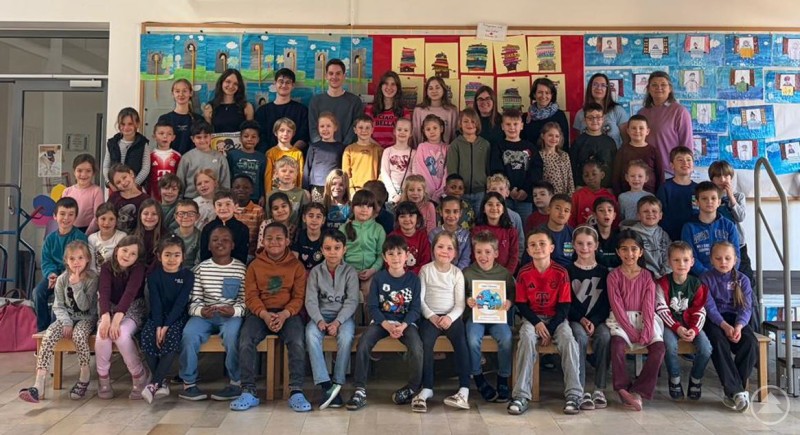 Eine gro&szlig;e Gruppe von Grundschulkindern sitzt und steht gemeinsam mit Lehrkr&auml;ften in einem Klassenzimmer vor bunt gestalteten W&auml;nden und pr&auml;sentiert stolz ein Buch.