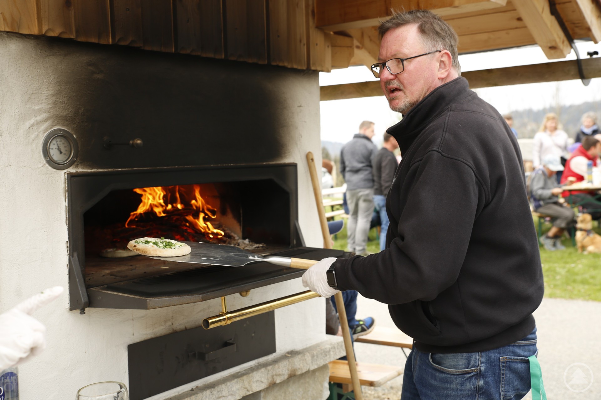 An der Kirche gab es Segen, am Backofen Sengzelten. Das eine für die Seele, das andere für den Magen. Das Bild zeigt einen Mann beim Backen von Sengzelten in einem traditionellen Holzofen. Mit Backschaufel und Handschuhen schiebt er die Fladen mit Kräutern in den Ofen, in dem bereits ein offenes Feuer lodert. Im Hintergrund sieht man Besucher, die auf Bierbänken sitzen und das gemütliche Beisammensein genießen.