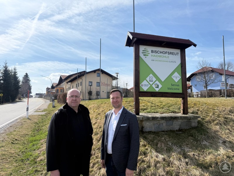 Bürgermeister Roland Schraml, Landtagsabgeordneter Josef Heisl. Das Bild zeigt zwei Männer, die vor einem großen Willkommensschild in Bischofsreut (Gemeinde Haidmühle) stehen. Das Schild trägt den Slogan „Am Grünen Band Europa“ und begrüßt Besucher mit „Servus / Welcome“. Im Hintergrund ist die Straße mit Häusern, darunter der „Witikohof“, zu sehen. Die Szene wirkt wie ein offizieller Besuch oder ein Pressetermin bei sonnigem Wetter in ländlicher Umgebung.
