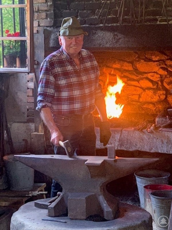 Ein Schmied in karierter Kleidung steht in einer traditionellen Schmiede vor einem lodernden Feuer und arbeitet an einem Amboss.