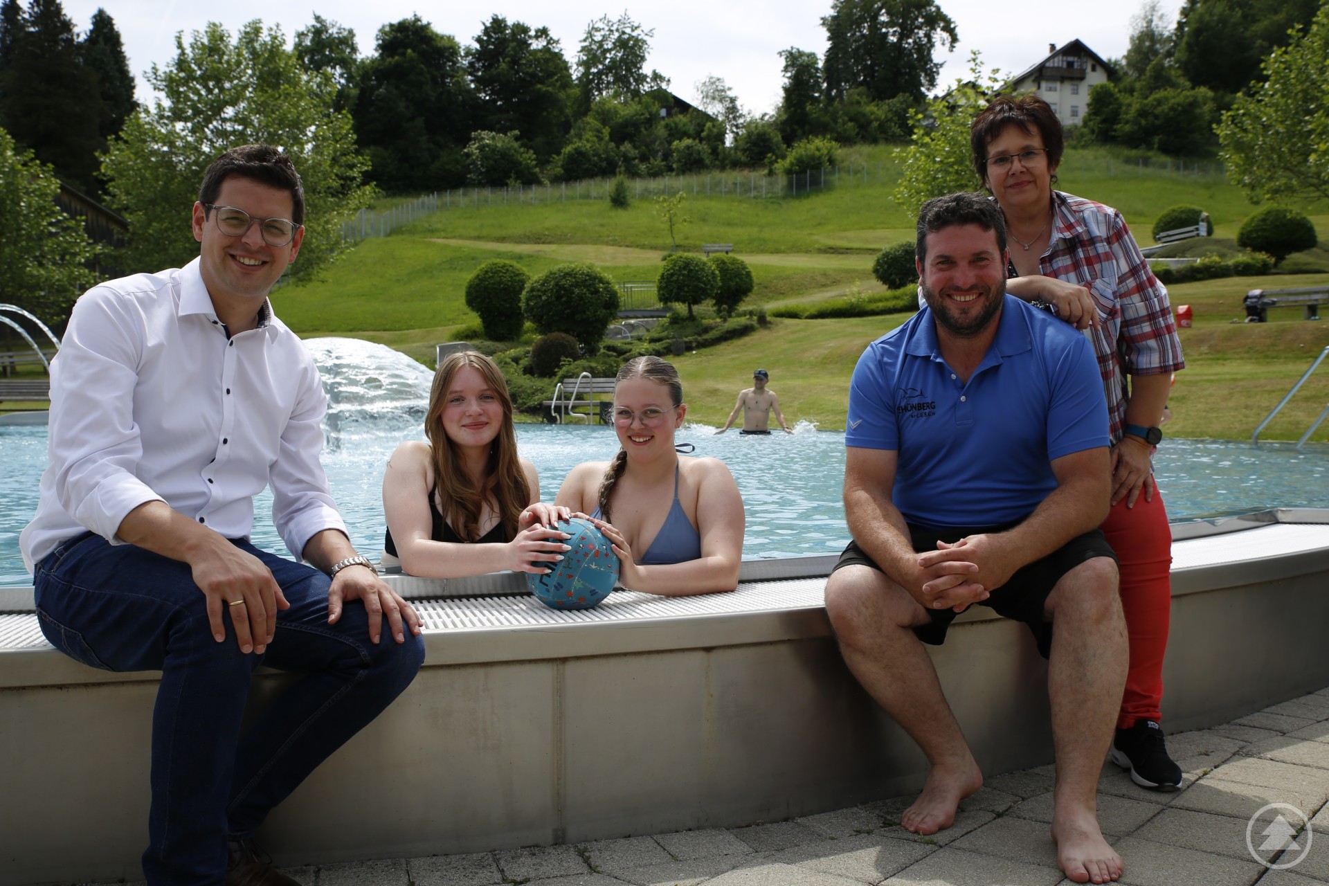 Alina und Theresa haben sich als Temperaturtester ganz zufrieden gezeigt mit der Startwärme im EBS. Die Sonne leuchtete schon mal kräftig zwischen vereinzelten Regenprognosen. Bürgermeister Martin Pichler (l.) sowie Kioskbetreiberin Conny Eder (v.r.) und Daniel Peschl als Bademeister freuen sich auf großen Andrang. Fünf Personen – zwei junge Frauen im Wasser und drei Erwachsene am Beckenrand – posieren lächelnd am Rand eines Schwimmbeckens im Erlebnisbad Schönberg. Im Hintergrund sind Liegewiese, Bäume und ein Haus auf einer Anhöhe zu sehen.