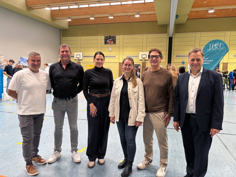 Sechs Personen stehen in einer Turnhalle vor Messeständen und lächeln in die Kamera.