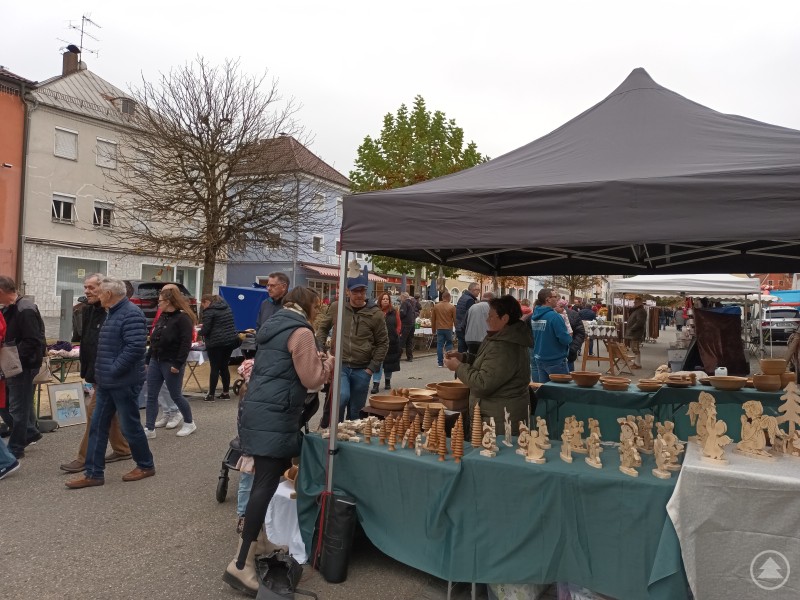 Menschen flanieren über den belebten Marktplatz, der mit Marktständen und Pavillons gefüllt ist. An einem Stand werden handgefertigte Holzfiguren und Schalen angeboten.