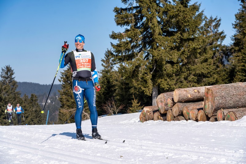 Skimarathon am Bretterschachten: Der Skadi Loppet zählt zu den schweren Strecken im europäischen Skimarathon. Ein Langläufer steht auf einer verschneiten Loipe, neben ihm liegen Holzstapel, im Hintergrund ragen Bäume in den blauen Himmel.