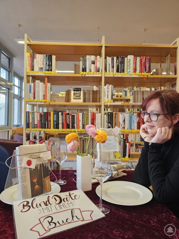 Überraschende Reisegeschichten erwarten die Besucher beim „Blind Date mit dem Buch“ in der Stadtbibliothek Deggendorf. Eine Frau sitzt an einem mit rotem Samttuch gedeckten Tisch in einer Bibliothek. Vor ihr stehen Weingläser, ein Teller, ein Blumenstrauß und mehrere verpackte Bücher mit Hinweisschildern, auf einem liegt ein Schild mit der Aufschrift „Blind Date mit einem Buch“. Im Hintergrund sind zahlreiche Bücherregale zu sehen.