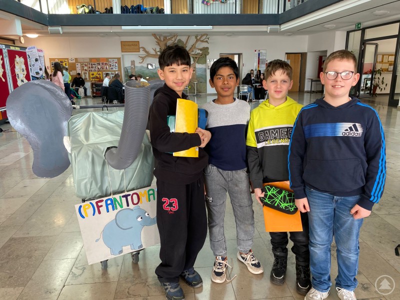 Schüler präsentieren den selbst gebauten (P)FANTOMAT, eine kreative Sammelstation für Pfandflaschen am LLG Grafenau. Vier Schüler stehen in der Aula des Landgraf Leuchtenberg Gymnasiums neben dem selbst gebauten (P)FANTOMAT. Die Sammelstation ist als grauer Elefant gestaltet und dient zum Einwerfen von Pfandflaschen.