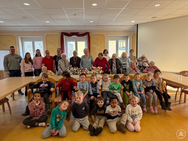 hinten von links nach rechts Einrichtungsleiter Gerald Schröder, Mitarbeiterinnen des Kindergartens und der Betreuung, Bewohnerinnen und Bewohner von St. Helena, 6. Von rechts PDL Frau Kormaier Brigitte, vorne Kinder des Caritas Kindergartens am Kirchplatz. Eine große Gruppe aus Senioren und Kindergartenkindern sitzt und steht gemeinsam in einem hellen Raum. Vor ihnen liegen viele bunt bemalte Ostereier, während alle in die Kamera schauen.