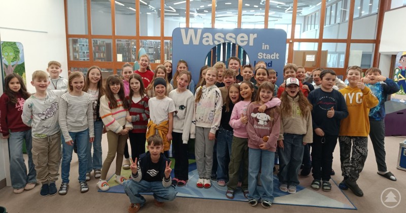 Klassen 4a und 4b auf der Suche nach Wasser in Stadt und Land. Eine große Gruppe von Grundschulkindern steht gemeinsam vor einer Ausstellung mit dem Titel Wasser in Stadt und Land. Die Kinder lächeln in die Kamera und befinden sich in einem hellen Raum mit Glaswänden und Ausstellungselementen.