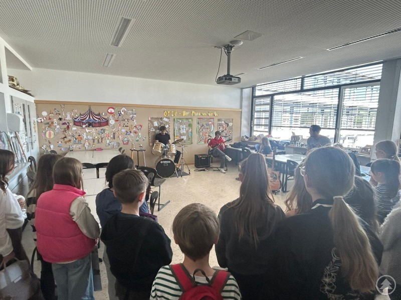 Sch&uuml;lerinnen und Sch&uuml;ler stehen in einem Musikraum vor einer Schulband mit Schlagzeug und Gitarre und h&ouml;ren der Musikdarbietung zu.