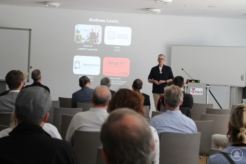 Andreas Lovric ging in der Keynote auf das Thema „Zukunft ist kein Zufall“ ein. Ein Mann spricht vor Publikum in einem Vortragssaal, im Hintergrund ist eine Präsentationsfolie mit seinem Namen und Tätigkeitsfeldern zu sehen.