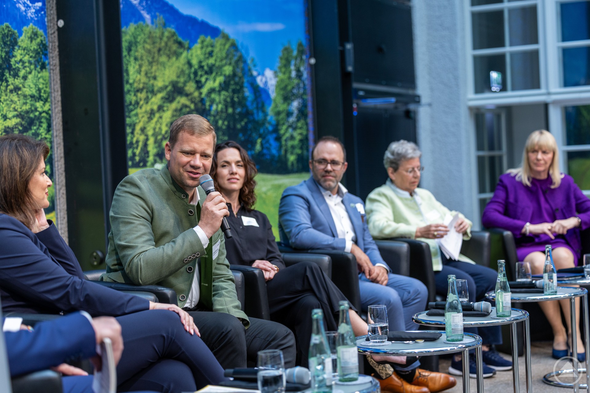 Podiumsdiskussion mit sechs Teilnehmenden, darunter Dr. Olaf Heinrich mit Mikrofon, im Innenbereich vor einem großformatigen Landschaftsbild.