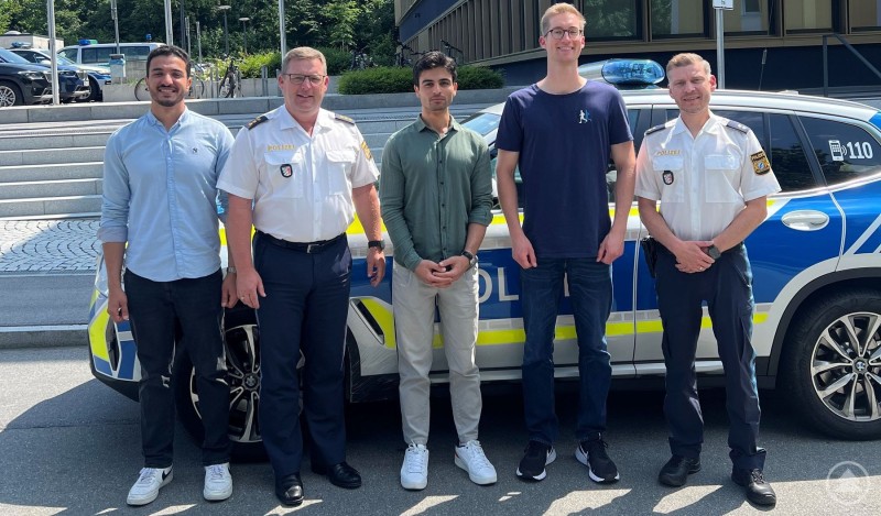 Polizei, v.l.n.r. Herr Mahmoud Hesham Abdelhakeem Elsayed Edree, Polizeidirektor Christian Dichtl, Herr Ramazan Bozkurt, Herr Fynn Brackmann, Polizeioberkommissar Johannes Weigerstorfer Fünf Männer stehen nebeneinander vor einem Polizeiauto. Zwei von ihnen tragen Polizeiuniformen, die anderen drei sind zivil gekleidet. Sie lächeln in die Kamera, im Hintergrund sind weitere Polizeifahrzeuge und ein modernes Gebäude zu sehen.