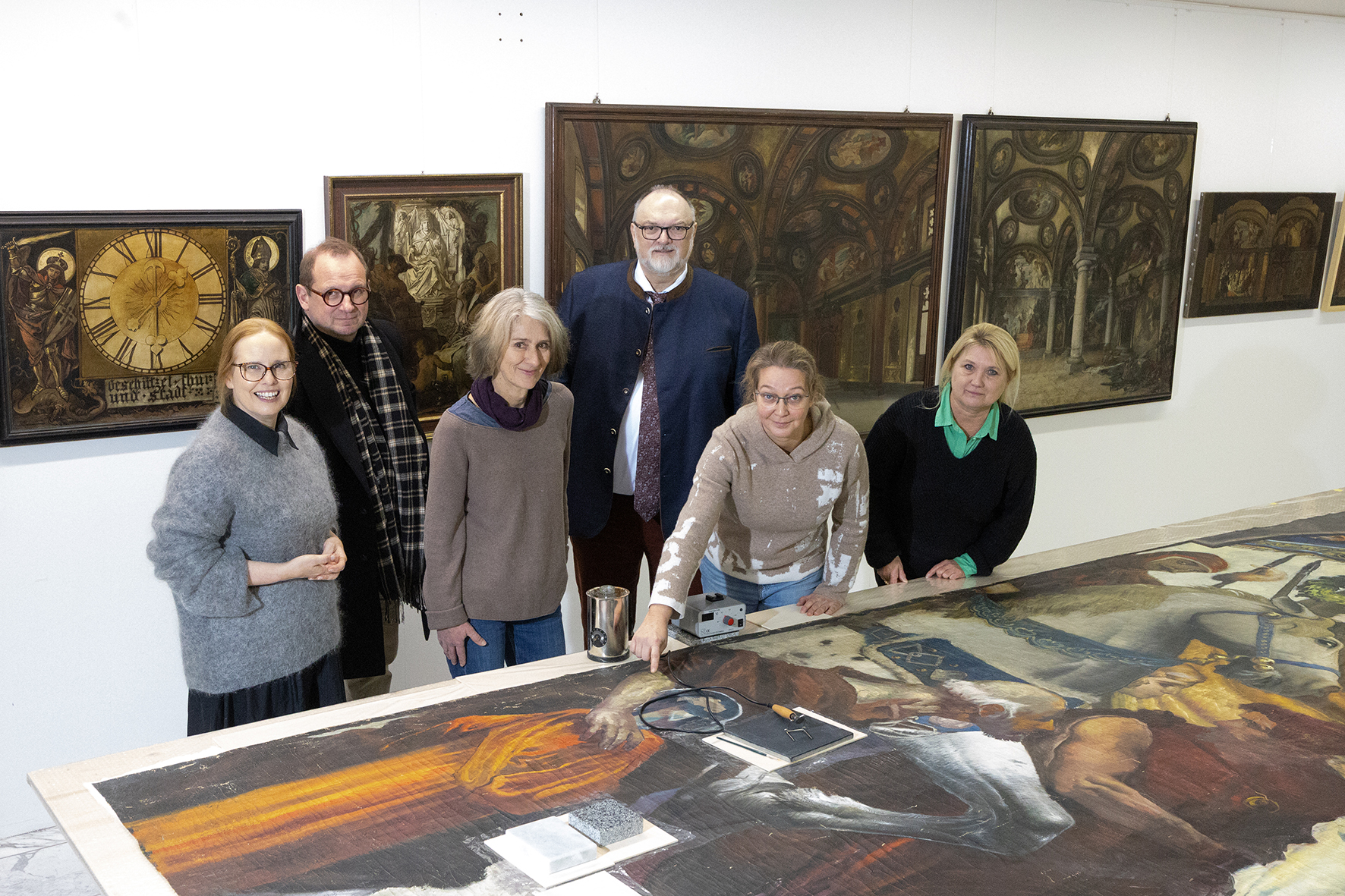 Sechs Personen stehen in einem Restaurierungsatelier hinter einem großformatigen Gemälde. Im Hintergrund hängen weitere historische Gemälde an der Wand.