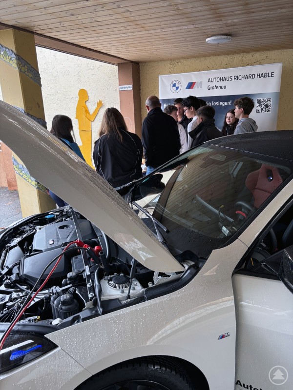 Interessiert arbeiten die Schülerinnen und Schüler am Testgerät zum Auslesen von Fehlern an einem Kraftfahrzeug. Eine Gruppe Jugendlicher steht in einer Werkstatt neben einem geöffneten Auto, dessen Motorraum sichtbar ist.