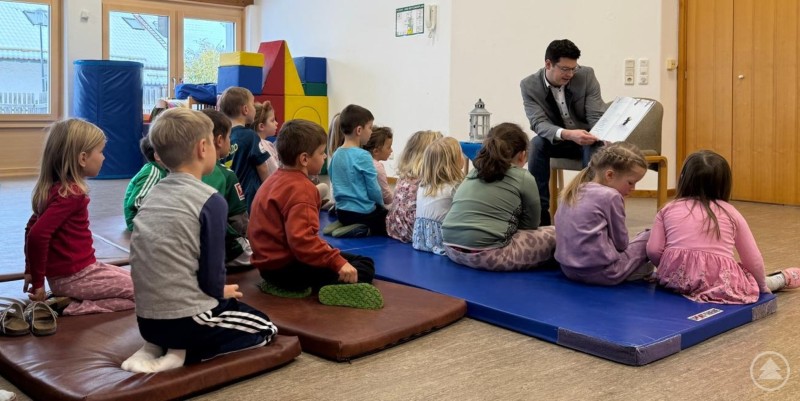 Jetzt muss sich Bürgermeister Martin Pichler wieder auf die Suche nach Abenteuern machen, womit er nächstes Jahr wieder Kinder mit Buchstaben fesseln kann. Mehrere Kindergartenkinder sitzen aufmerksam auf Matten und Kissen in einem Gruppenraum, während ein Mann im Anzug – Bürgermeister Martin Pichler – ihnen aus einem Bilderbuch vorliest. Die Kinder sind konzentriert und gespannt, das Ambiente ist freundlich und gemütlich.