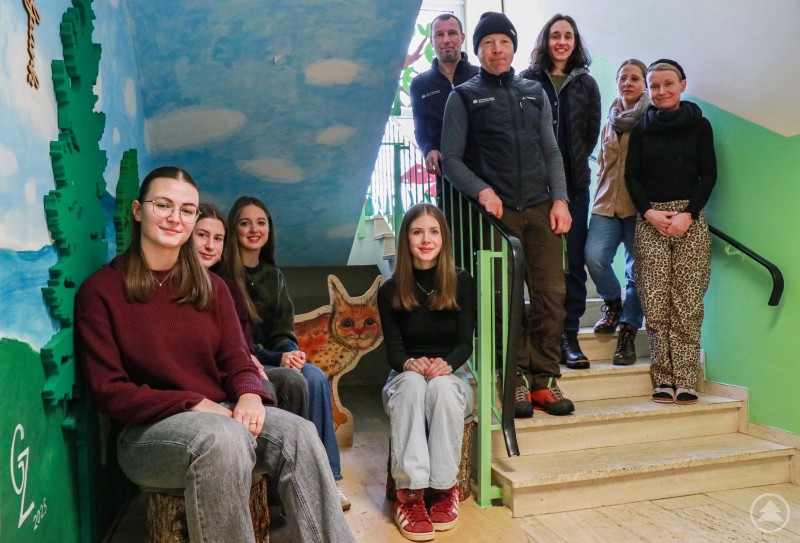Die vier Gymnasiastinnen Mia Probst, Ronja Obermeier, Elina Aichele und Marina Baierl (links) sowie die Haustechniker Max Schwarz und Martin Weber gestalteten die neue Nationalpark-Ecke im AWO-Kinderhaus in Zwiesel, die nun offiziell eröffnet wurde. Eine Gruppe aus vier jungen Frauen und fünf Erwachsenen steht und sitzt in einem freundlich bemalten Treppenhaus mit Nationalpark-Motiven. Die Schülerinnen sitzen auf Baumstumpfhockern, im Hintergrund sind bemalte Bäume und ein Luchs zu sehen.