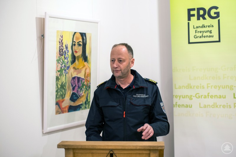 Kreisbrandrat Martin Bauer richtete ein Grußwort an die Geehrten. Kreisbrandrat Martin Bauer steht am Rednerpult und spricht vor der FRG Ehrenamt Wand.