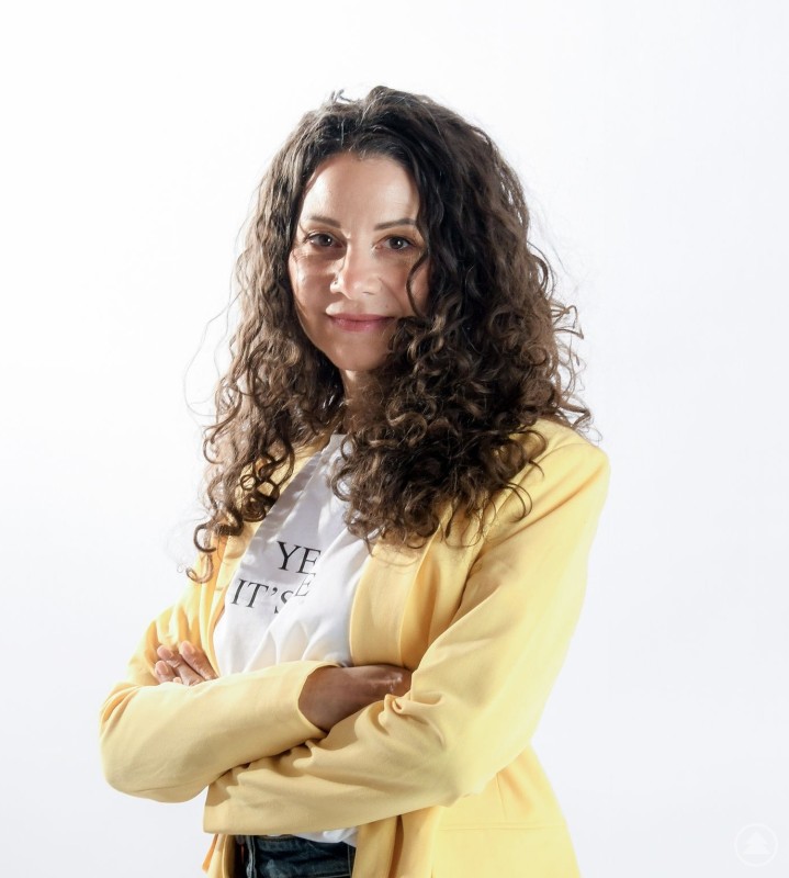 Birgit Weinert aus Hohenau engagiert sich als HPV Botschafterin für mehr Aufklärung und Prävention in der Region. Porträt einer Frau mit lockigem, schulterlangem Haar, die selbstbewusst mit verschränkten Armen vor hellem Hintergrund steht und einen gelben Blazer über einem weißen Shirt trägt.