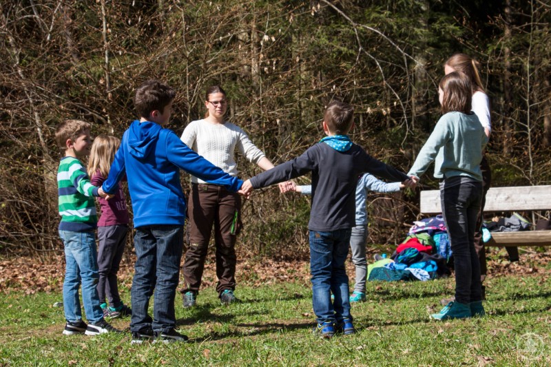 Spannende Outdoorerlebnisse warten auf die Kinder in den Osterferien, wie etwa im Waldspielgelände Spiegelau. Eine Gruppe von Kindern steht im Kreis auf einer Wiese im Wald und hält sich an den Händen, während eine Betreuerin in der Mitte steht und das Spiel anleitet.