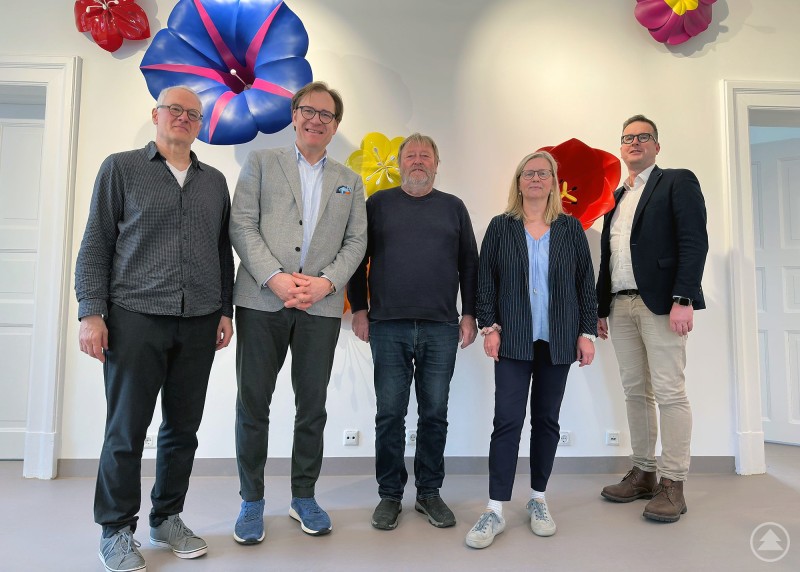 Im Bild v. l.: Daniel J. Schreiber, Bezirkstagsvizepräsident Dr. Thomas Pröckl, Hubert Huber, Anette Röhr und Bezirksheimatpfleger Dr. Clemens Knobling. Fünf Personen stehen in einem hellen Raum vor großen farbigen Blumenobjekten an der Wand und blicken in die Kamera.