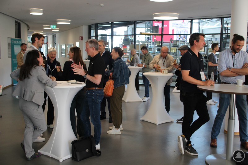 Menschen stehen in kleinen Gruppen an Stehtischen in einem modernen Foyer und unterhalten sich angeregt bei Getränken und Snacks.