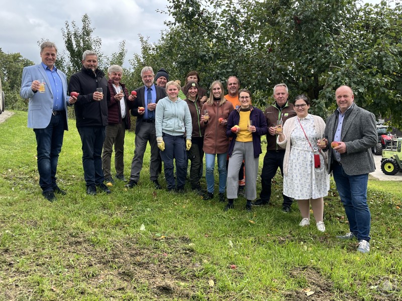 Die Streuobstaktion, mit dabei v.l.: Alexander Mayer (1. BGM Grafenau), Erwin Braumandl (1. BGM Neukirchen vorm Wald), Leo Meier (1. BGM Röhrnbach), Uwe Urtel (2. BGM Tiefenbach), Hans Kellermann, Melissa Schindler, Ute Ellwein, Christina Uhrmann, Corina Molz (Geschäftsführerin ILE Ilzer Land), Rupert Veit (Bauhofleiter Ruderting), Pia Auberger (Projektmanagerin Öko-Modellregion Passauer Oberland), Thomas Lehner (Leiter Staatsgut Kringell), Carolin Pecho (1. BGM Ringelai), Max Rosenberger (1. BGM Hutthurm). Eine Gruppe von dreizehn Personen steht auf einer Wiese vor Apfelbäumen, hält Äpfel und Gläser mit Apfelsaft in der Hand und lächelt in die Kamera.