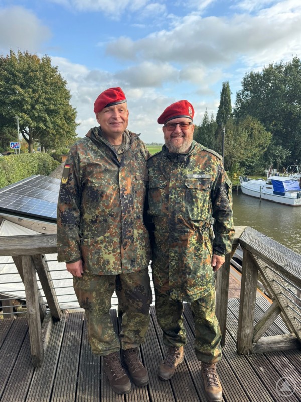Zwei Soldaten in Uniform mit roten Baretten lächeln in die Kamera, im Hintergrund Boote und Wasser