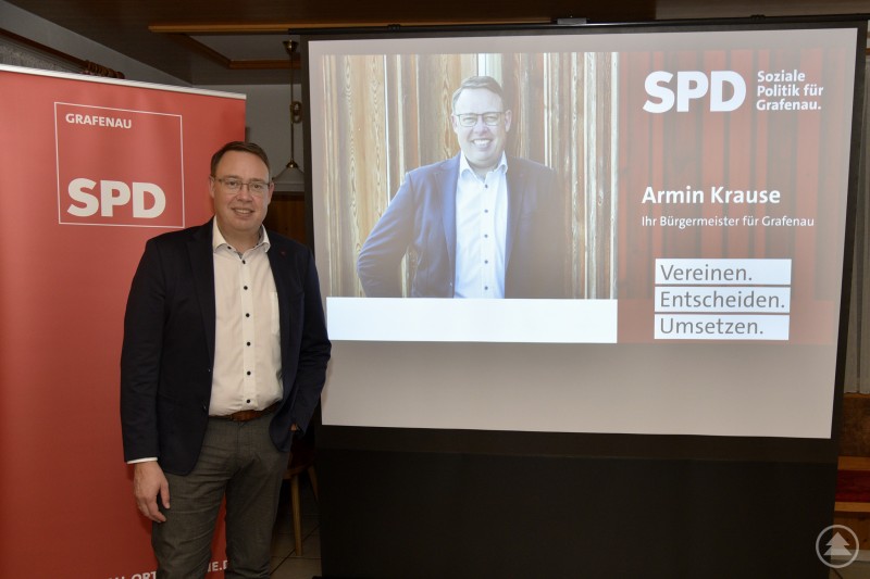 Ein Mann steht vor einem roten SPD-Rollup mit der Aufschrift „SPD Grafenau“. Daneben wird auf einer Leinwand ein Foto des gleichen Mannes mit den Worten „SPD – Soziale Politik für Grafenau. Armin Krause – Ihr Bürgermeister für Grafenau. Vereinen. Entscheiden. Umsetzen.“ präsentiert.