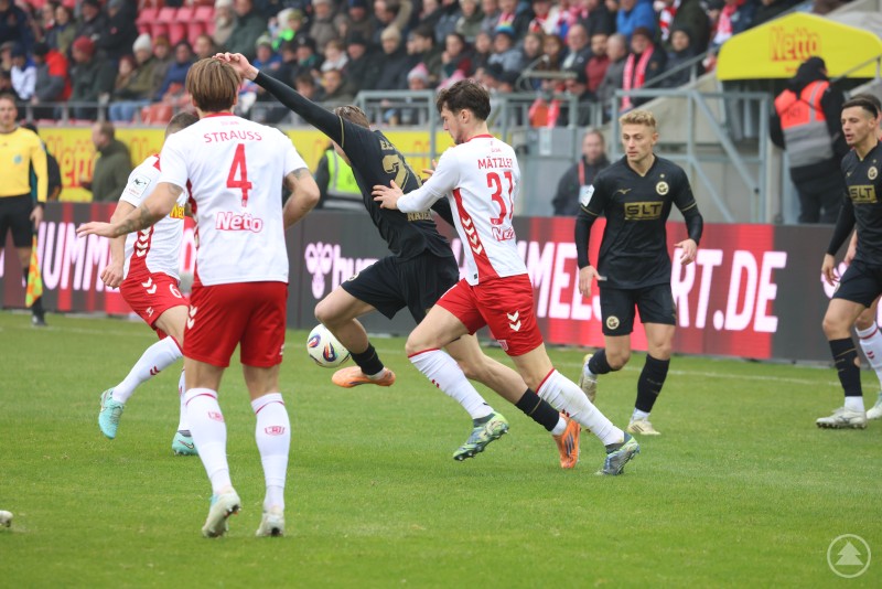 Intensiver Zweikampf zwischen Jahn Regensburg und Hansa Rostock während des Spiels im Jahnstadion. Zwei Spieler des SSV Jahn Regensburg (weiße Trikots, rote Hosen) im Zweikampf mit einem Spieler von Hansa Rostock (schwarzes Trikot). Der Ball befindet sich zwischen den Spielern, im Hintergrund sind weitere Akteure und Zuschauer auf der Tribüne zu sehen.